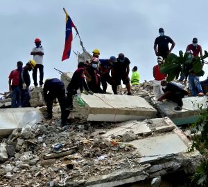 Rescatistas y voluntarios sobre los escombros de un edificio colapsado tras los terremotos del 24J, con una bandera de Venezuela